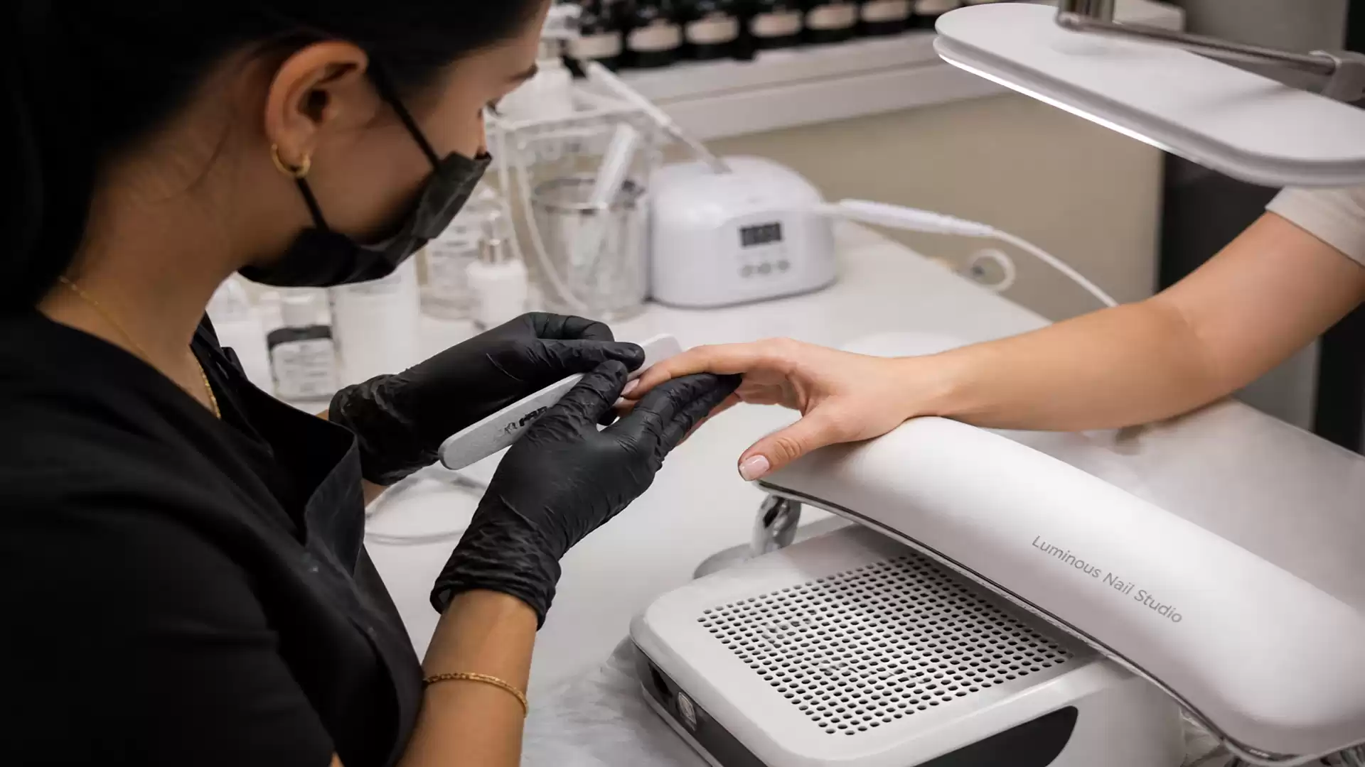 A nail technician wearing a black mask and black gloves files a customer's nails in a modern nail studio.