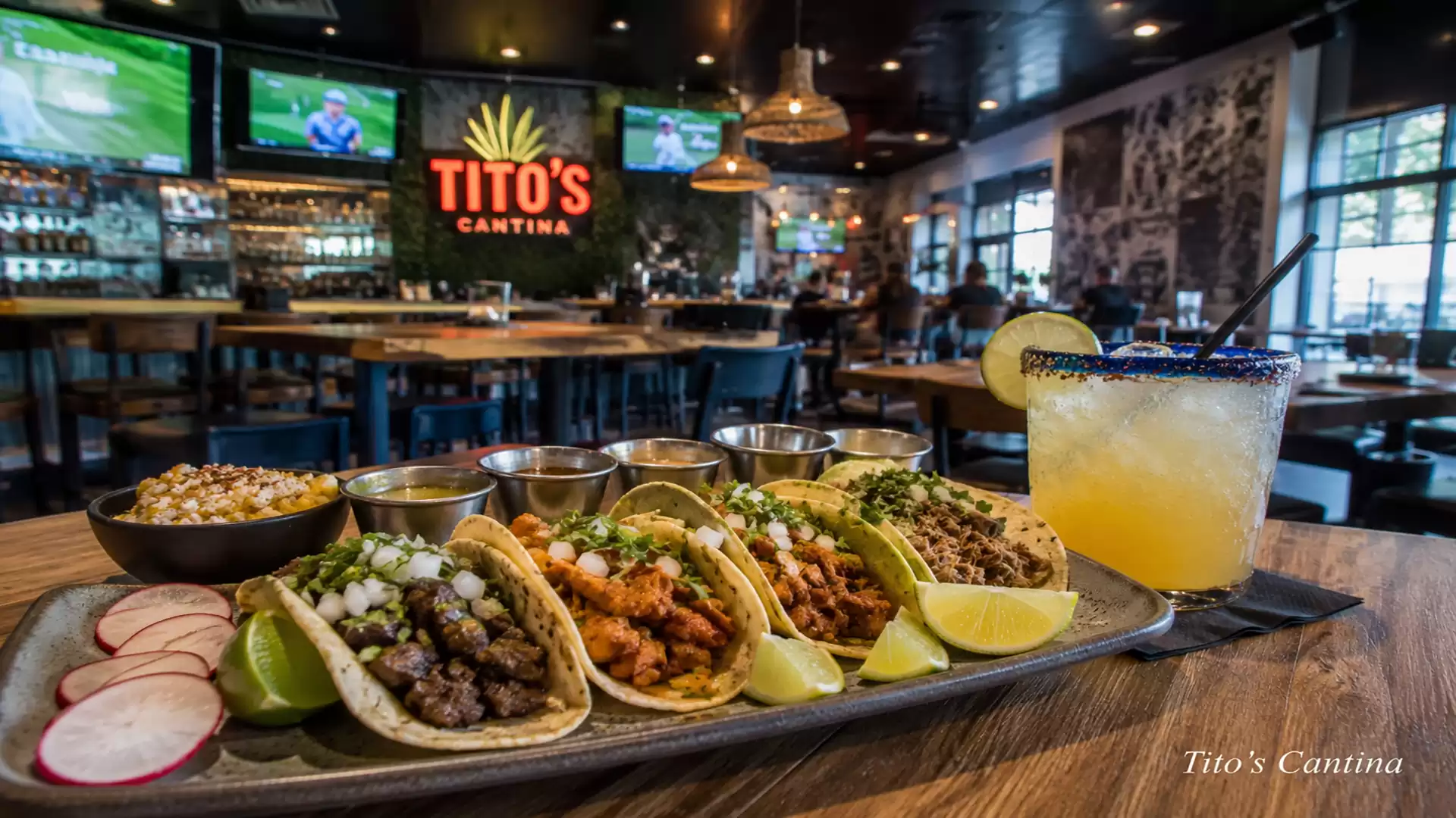 A stylish restaurant featuring a platter of five tacos garnished with onions and herbs, served alongside lime wedges. In the background, a cozy bar atmosphere at Tito’s Cantina, suitably lit, with a glass of orange cocktail with a lime slice and salted ri