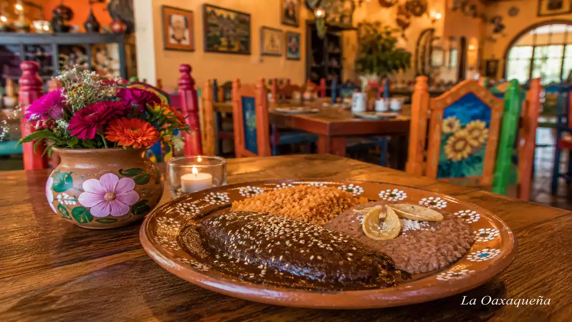 A traditional Mexican restaurant with colorful decor. In the foreground, a plate with mole, rice, and beans, next to a vase with flowers.
