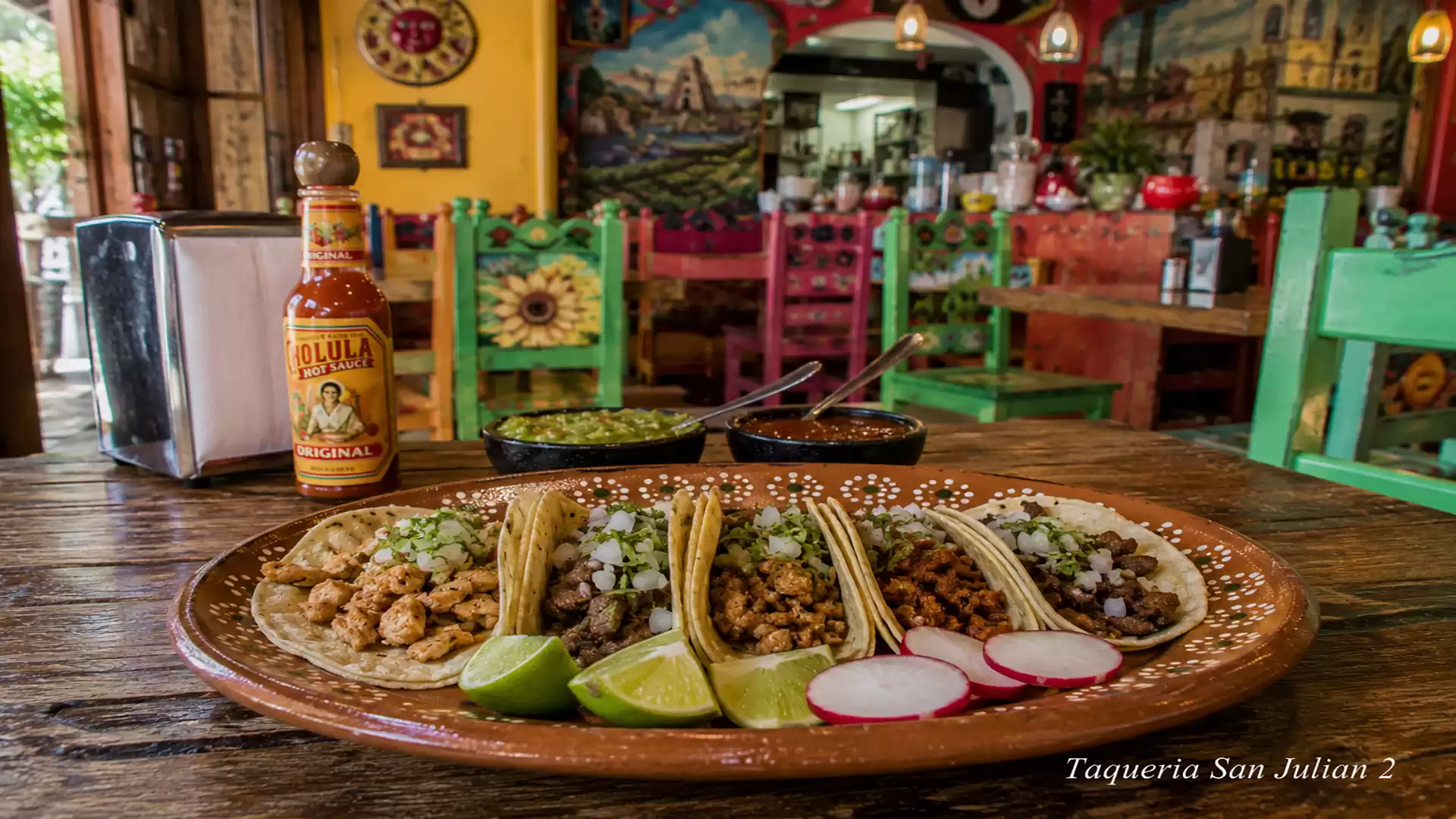 A plate of tacos in a brightly decorated restaurant. The table is adorned with a bottle of hot sauce and two small bowls of side dishes, surrounded by vibrant murals and colorful chairs.
