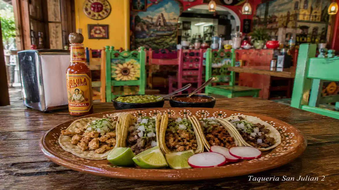 A plate of tacos in a brightly decorated restaurant. The table is adorned with a bottle of hot sauce and two small bowls of side dishes, surrounded by vibrant murals and colorful chairs.