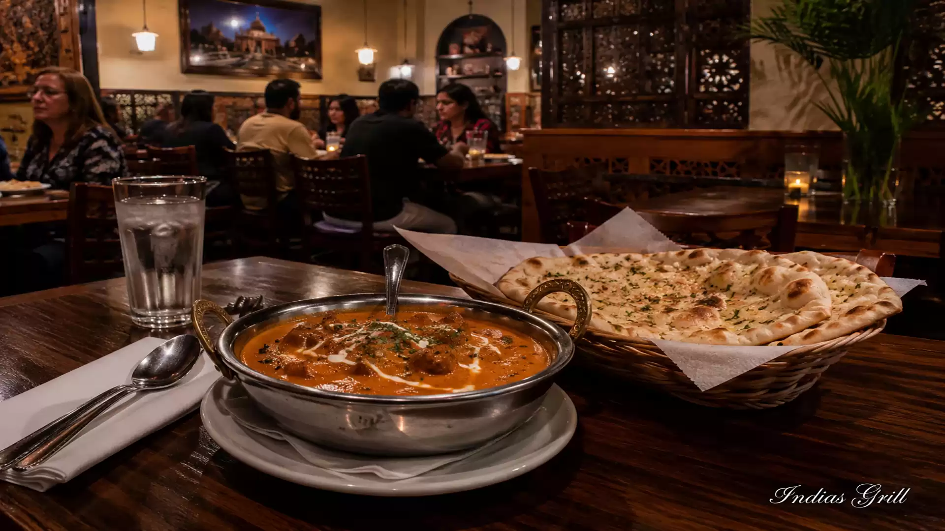 A cozy Indian restaurant with a bowl of curry and a basket of naan bread on a table. Guests are seated at tables in the background.