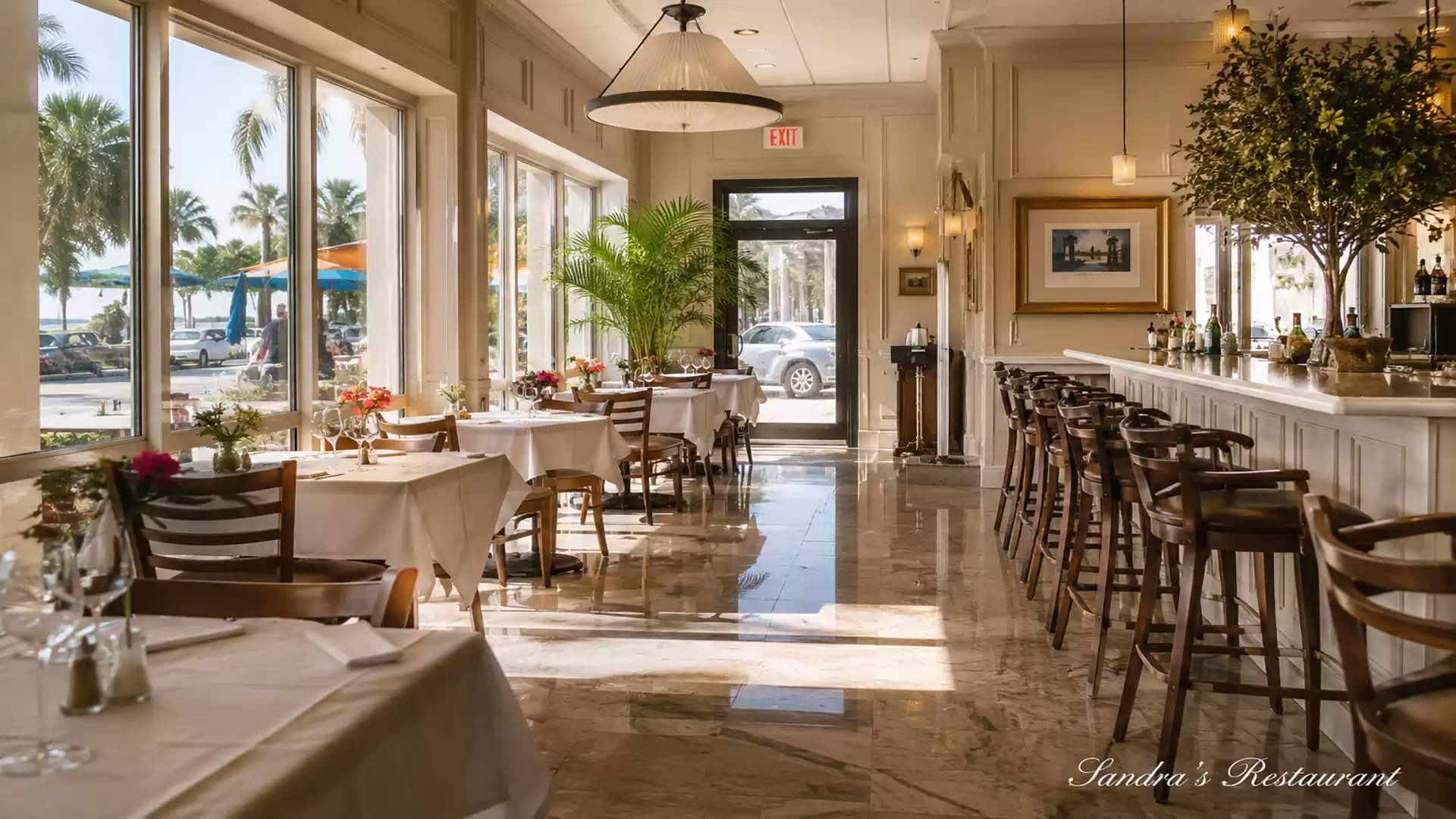 Beautiful restaurant with large windows letting in plenty of light, white tablecloths and wooden chairs, and a long bar with bar stools.