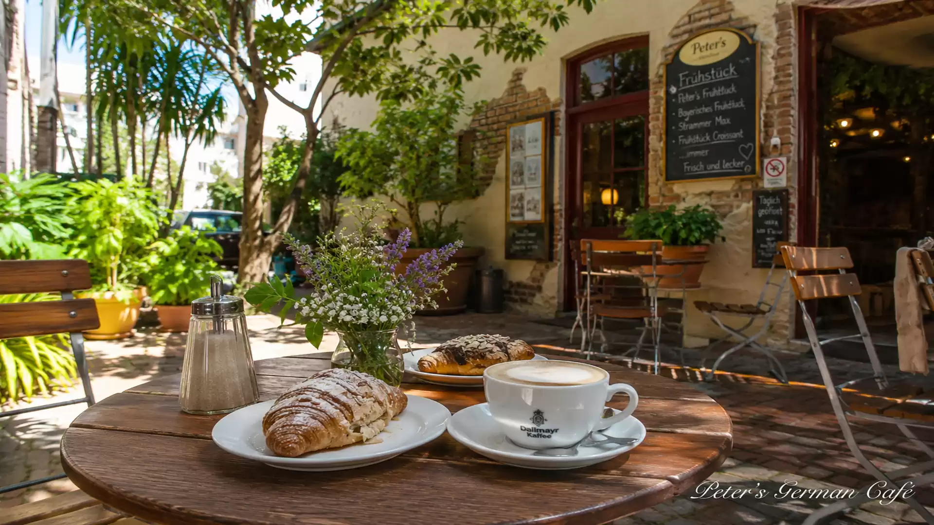 A cozy outdoor café with a round wooden table holding a croissant, a piece of cake, and a cappuccino. In the background, there's a charming building with a chalkboard menu and lots of plants.
