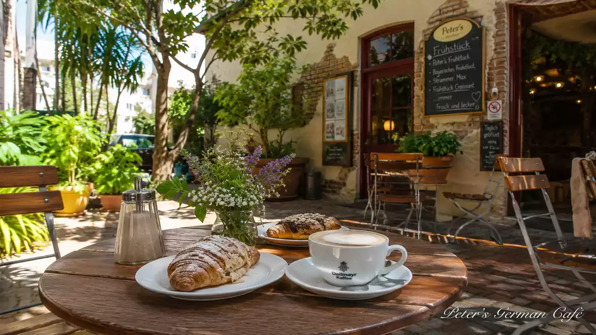 A cozy outdoor café with a round wooden table holding a croissant, a piece of cake, and a cappuccino. In the background, there's a charming building with a chalkboard menu and lots of plants.