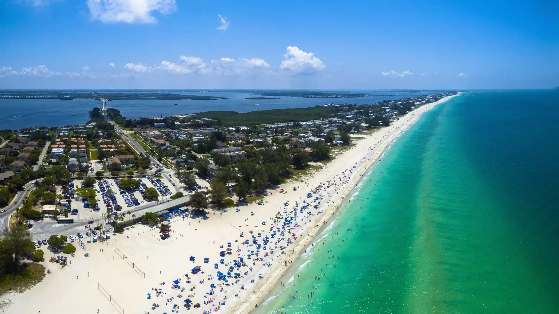 Aerial view of a busy beach with turquoise water and clear blue sky, many people and colorful umbrellas are visible.