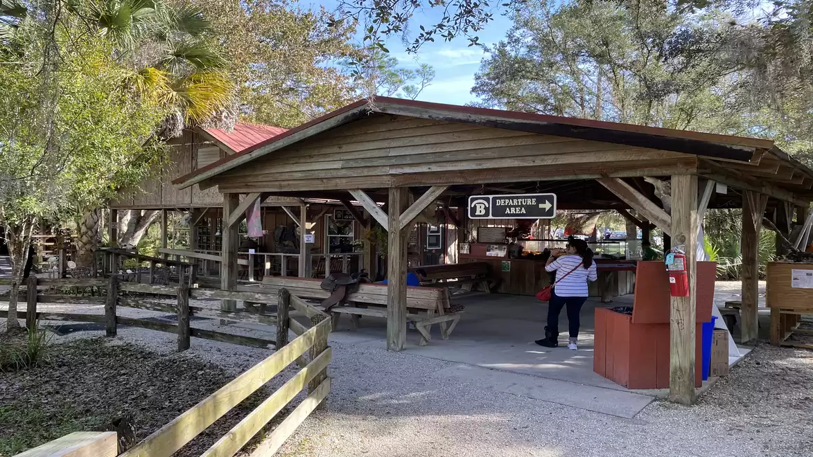 An outdoor covered wooden beer garden area with shaded trees in the background. A couple stands near a red trash bin.