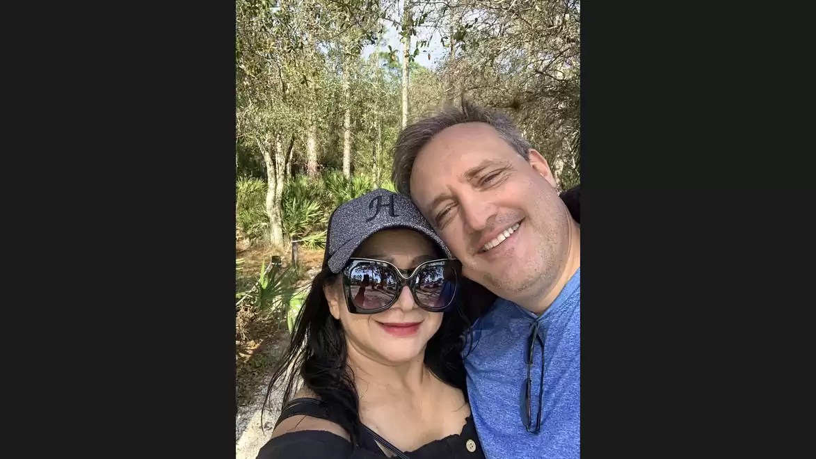 A smiling couple takes a selfie outdoors with trees in the background.