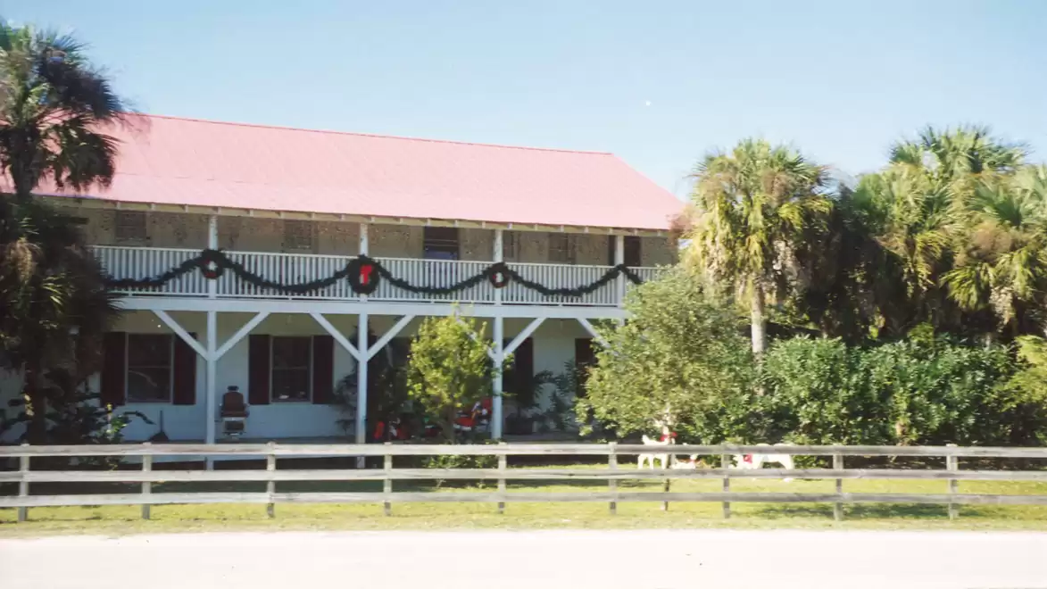 A two-story building with a red roof, surrounded by palm trees and bushes, decorated with garlands and a fence in front.