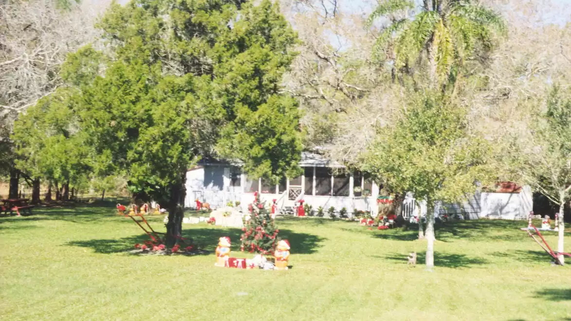 A white house with a porch surrounded by green trees and lawn. Christmas decorations like inflatable Christmas figures are scattered around the garden.
