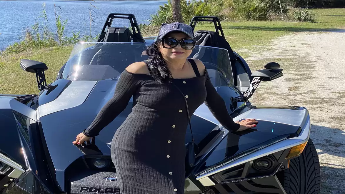 Woman in a black dress and sun hat standing in front of a black three-wheeled vehicle near a lake.