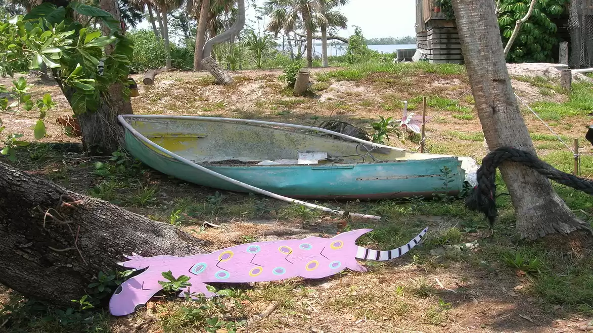 An old green rowboat lies on grass next to a tree trunk. In the foreground, there is a colorful, pink wooden figure on the ground. In the background, palm trees and a body of water are visible.