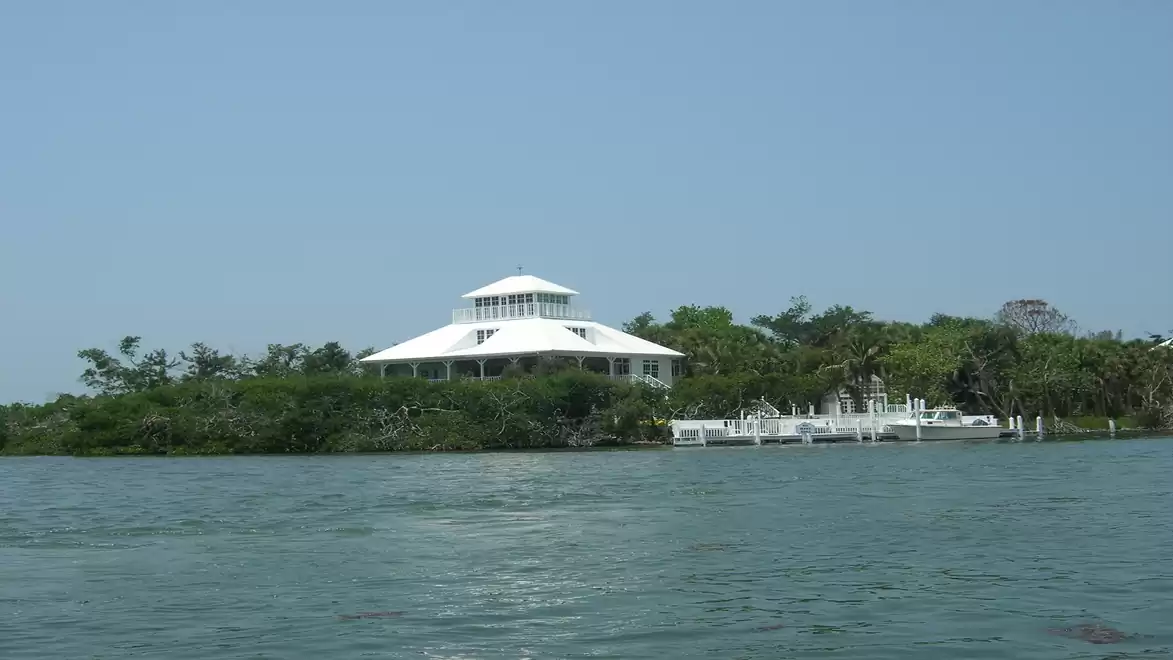 Large white house by the water with lush green vegetation in the background, sunny sky.