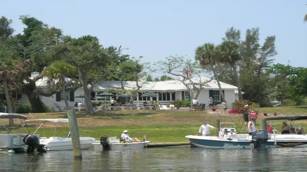 A white house with a porch by the water, surrounded by trees and boats in the foreground on a calm lake.