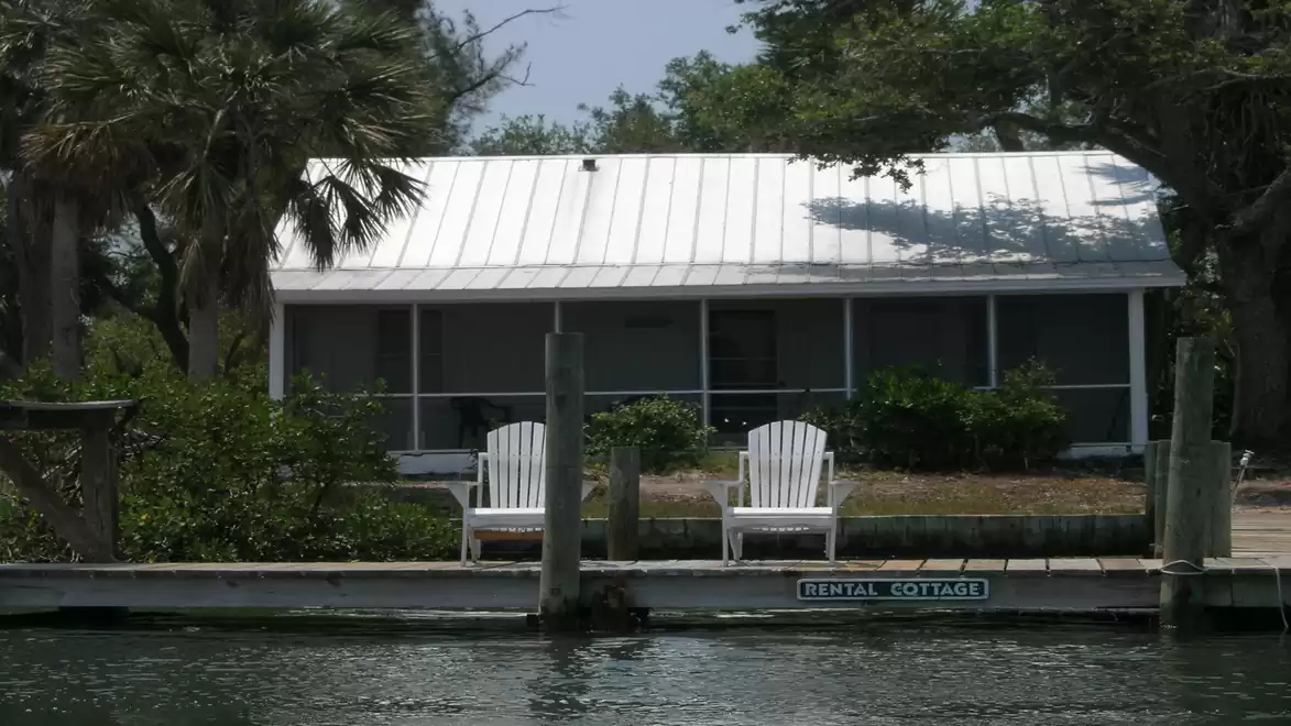 A charming white vacation cottage by the water with two white lounge chairs in front, surrounded by lush vegetation.