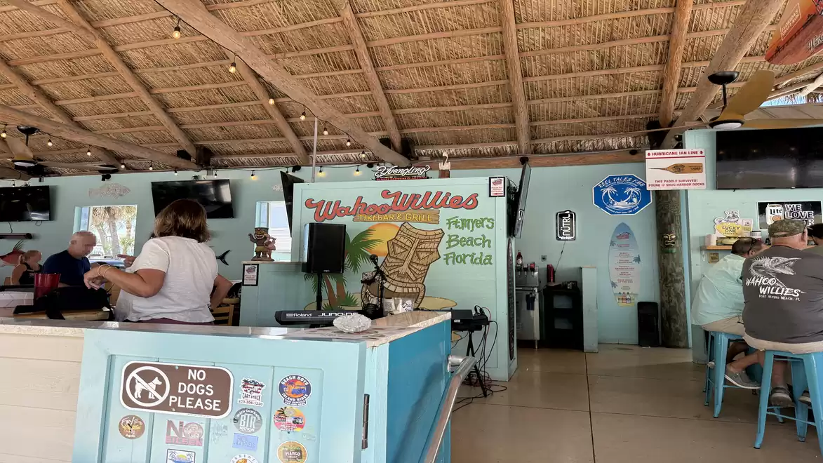 Interior view of a beach café with a thatched roof, colorful wall murals, and several people sitting at tables.