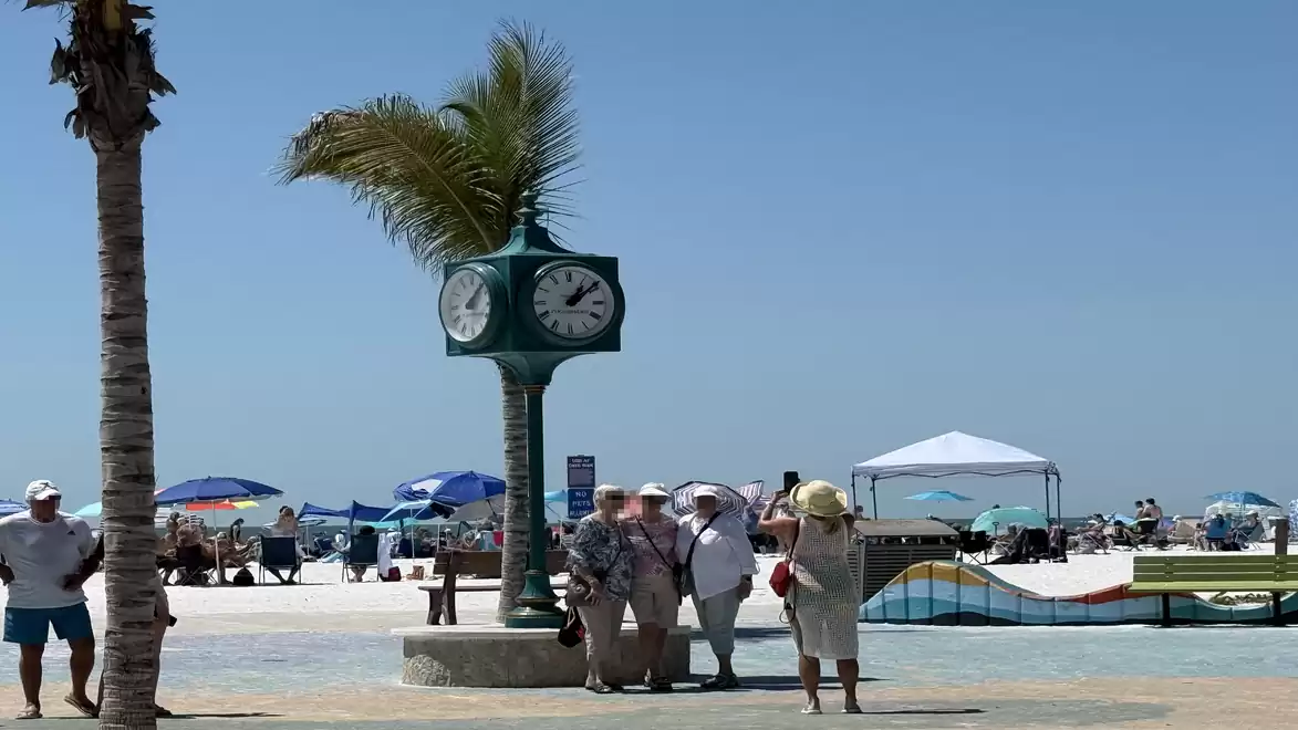Beach scene with a clock and palm trees, people strolling on the promenade, beach in the background with umbrellas.
