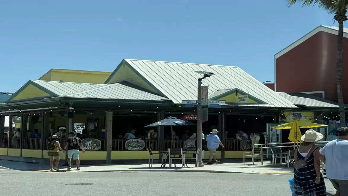A busy restaurant with a green roof structure under a blue sky. People are standing and sitting outside while some walk by.