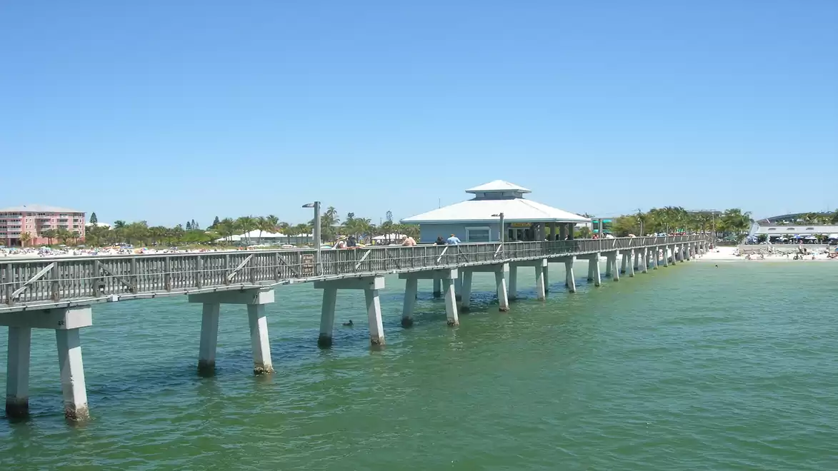 Long wooden pier on blue sea with pavilion, surrounded by sandy beach and green trees, under clear sky.