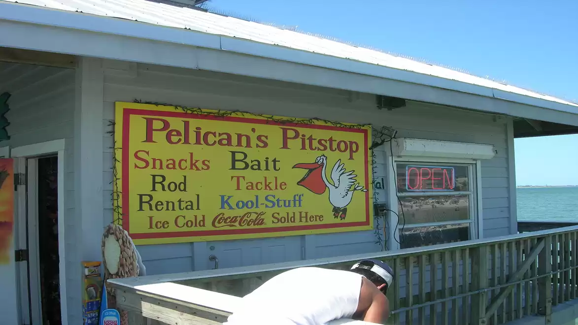 A sign on a wooden porch reads 'Pelican's Pitstop', offering items like snacks, bait, tackle, and rod rental. An 'Open' sign is visible in the window, and a person is wearing a white shirt.