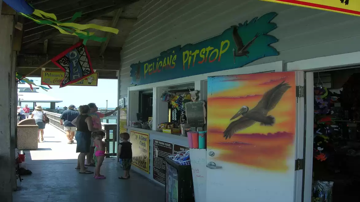 A shop on a pier with a painted pelican on the door. Customers are standing near the entrance as colorful flags flutter above the covered area.