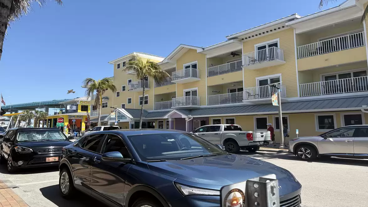 Street scene with parked cars in front of a yellow building with balconies and palm trees in the background.
