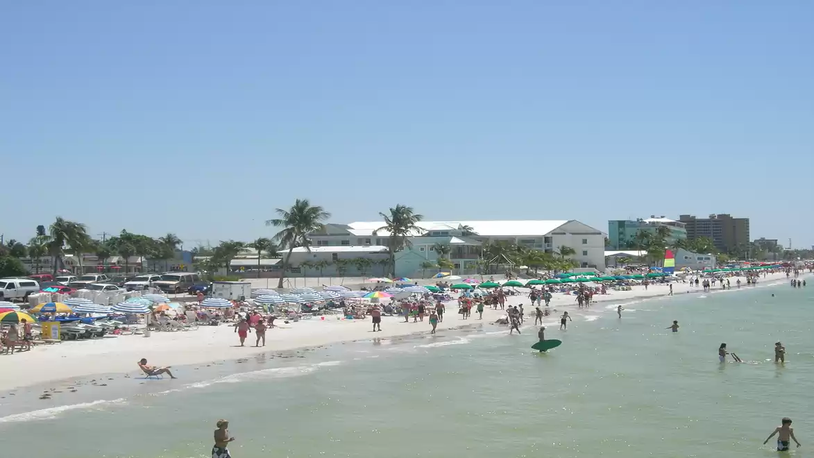Beach with many people in sunny weather, with palm trees and buildings in the background.