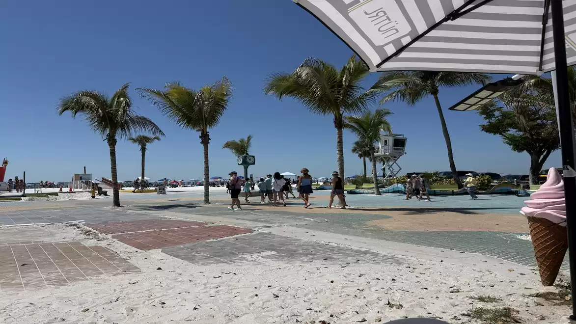 Sunny beach with palm trees, umbrella, and an ice cream cone in the foreground. People are walking along the promenade under the blue sky.
