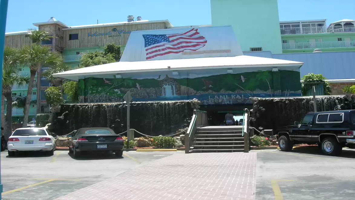 A tropical-style building with an American flag on the roof. Several cars are parked in front, and there are stairs leading to the entrance.