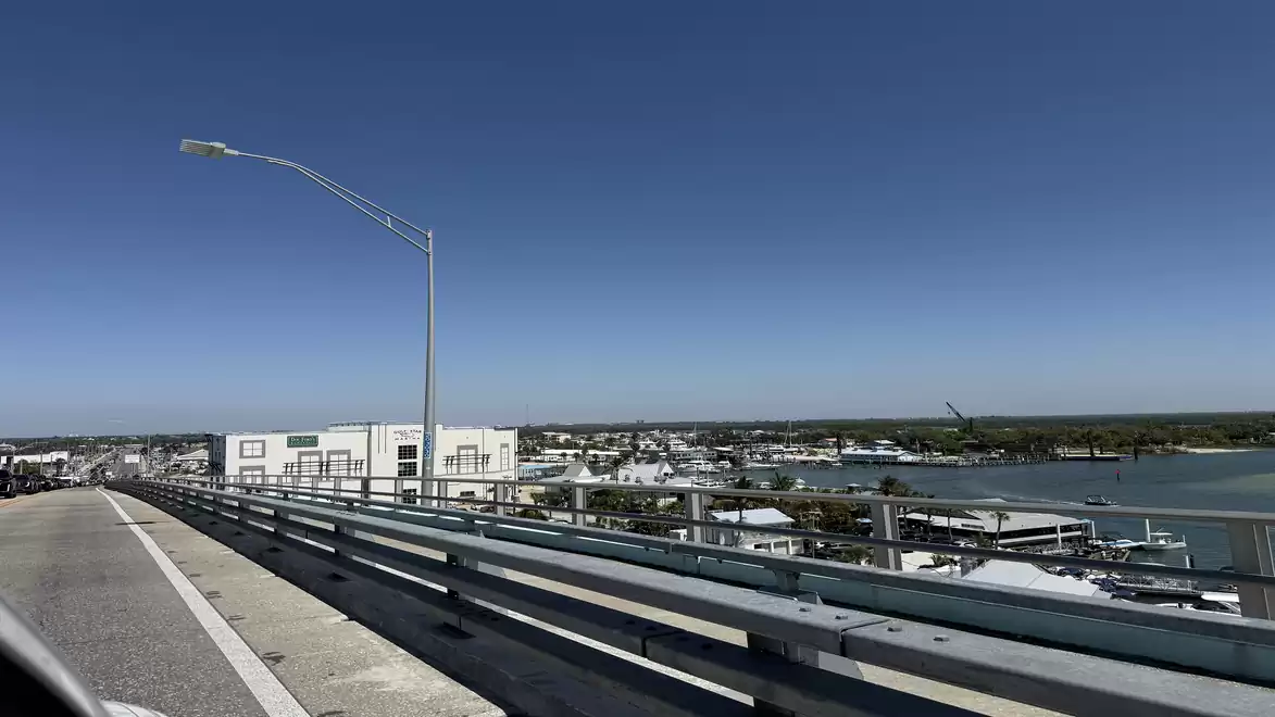 View of a bridge with a railing and a river in the background, flanked by buildings under a clear blue sky.