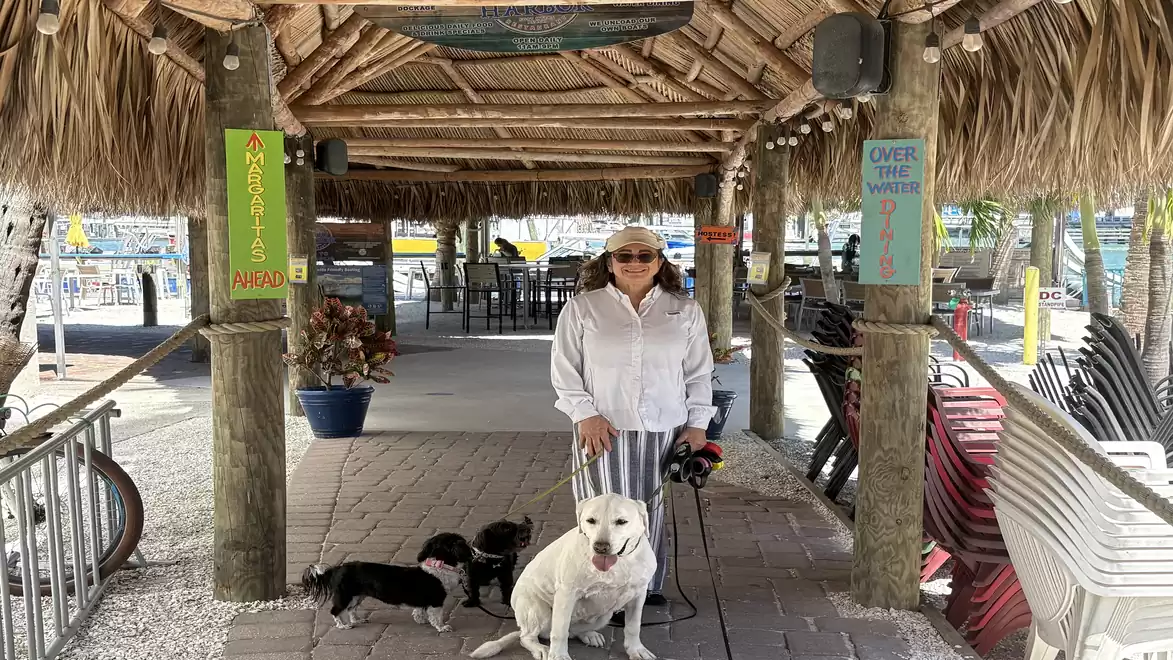 A woman stands under a thatched roof with two dogs, one sitting while the other stands.