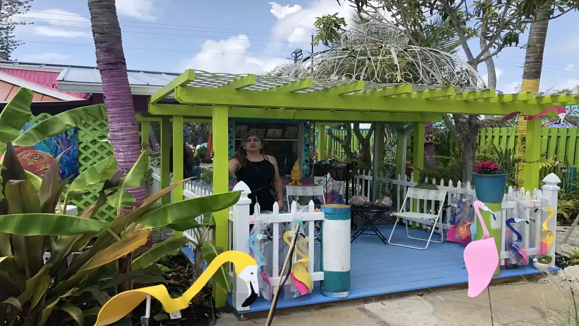 A colorful garden shed with a green roof, surrounded by plants and colorful decorations. A person stands in the background.