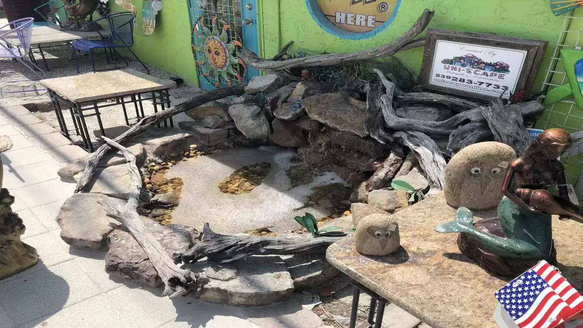 An outdoor area with a small fountain surrounded by natural stones and driftwood. There are decorative sculptures and a green-painted building in the background.
