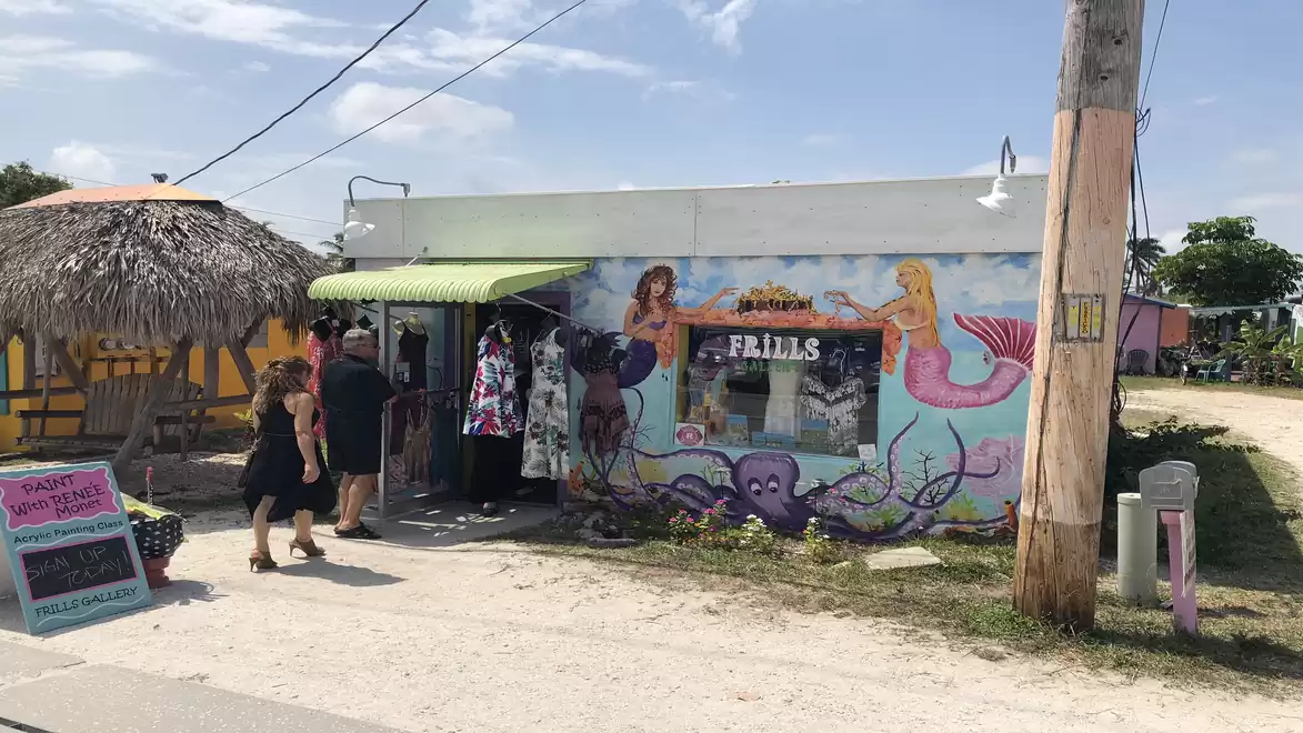 A small shop with colorful murals of mermaids and an octopus, tourists standing in front on a dusty road.
