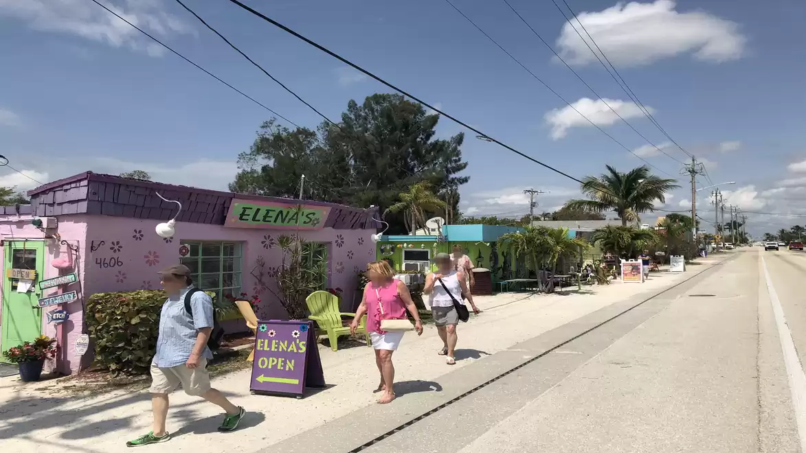 Street scene with pedestrians in front of a purple building called 'Elena's', surrounded by palm trees under a blue sky.