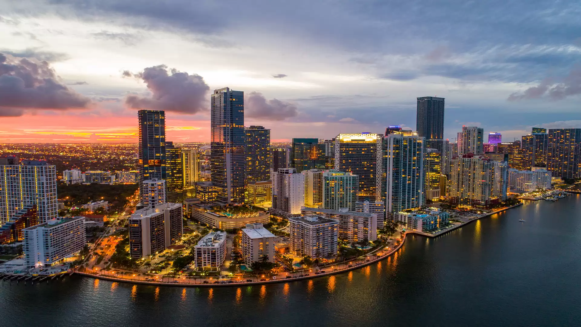 Panoramic view of Miami skyline at sunset, with illuminated skyscrapers by the water.