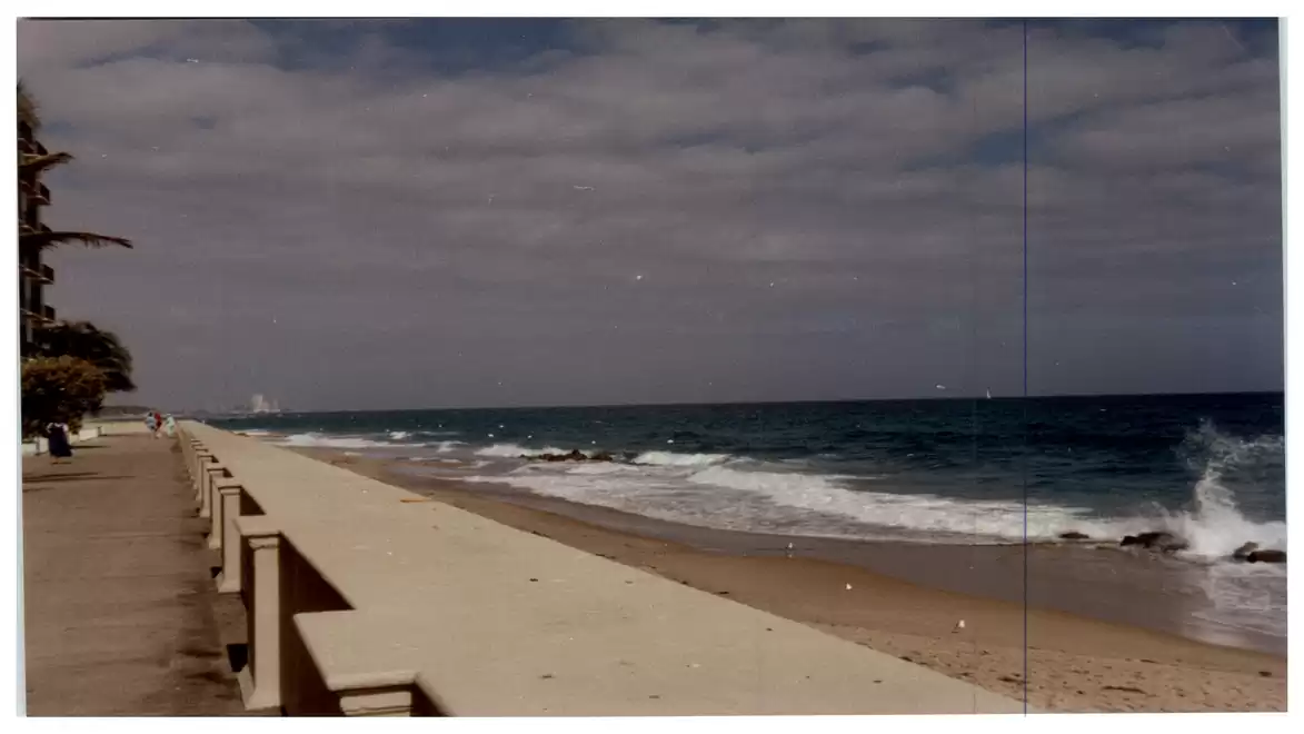 A beach with a concrete wall in the foreground, next to a calm ocean under a cloudy sky.