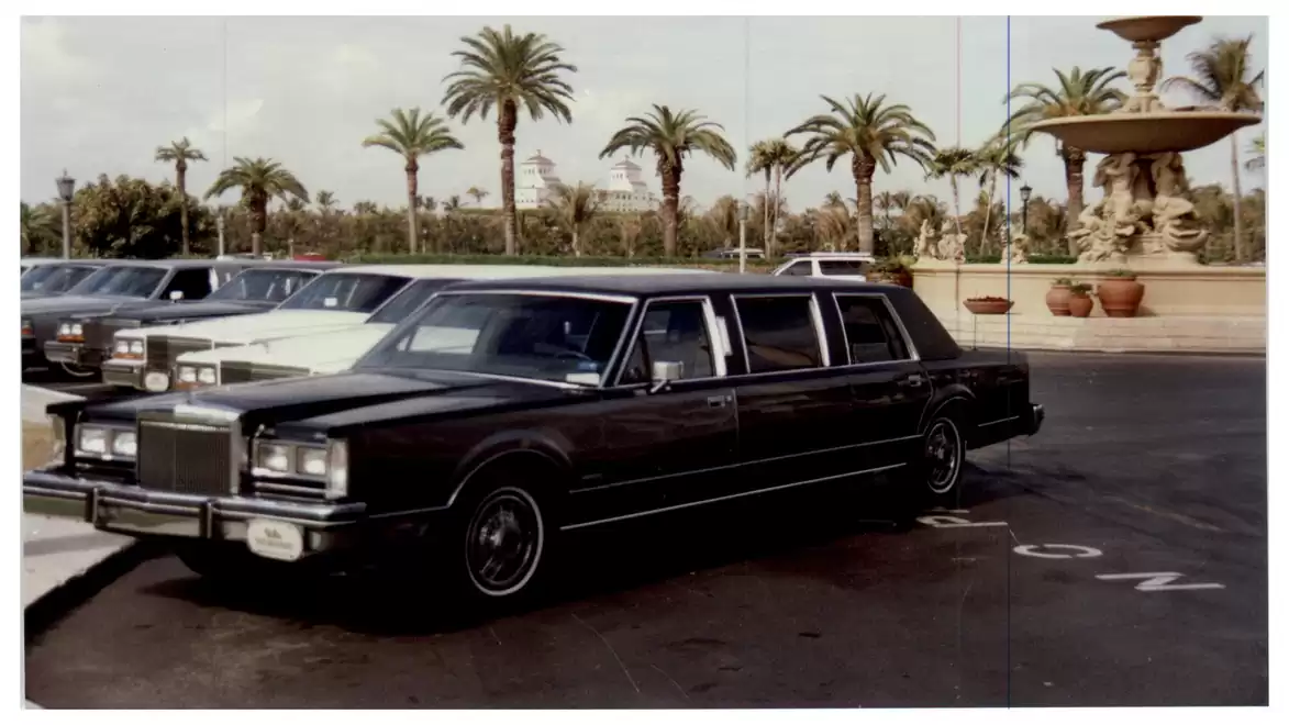 A black luxury car parked outdoors beside a tall, ornate water fountain and palm trees.