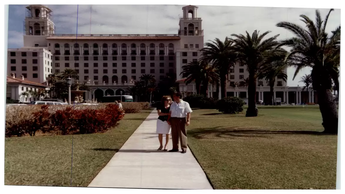 A couple walks along a pathway with a large hotel building and palm trees in the background.