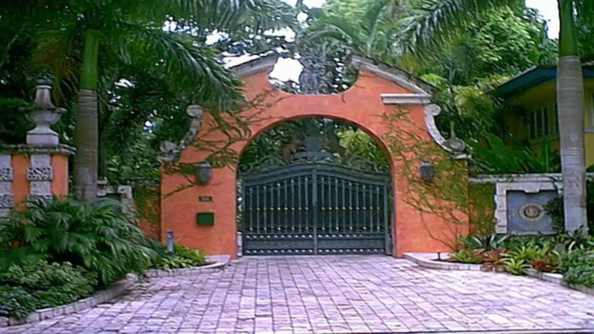 A large, ornate iron gate in front of an orange wall, surrounded by lush tropical plants.