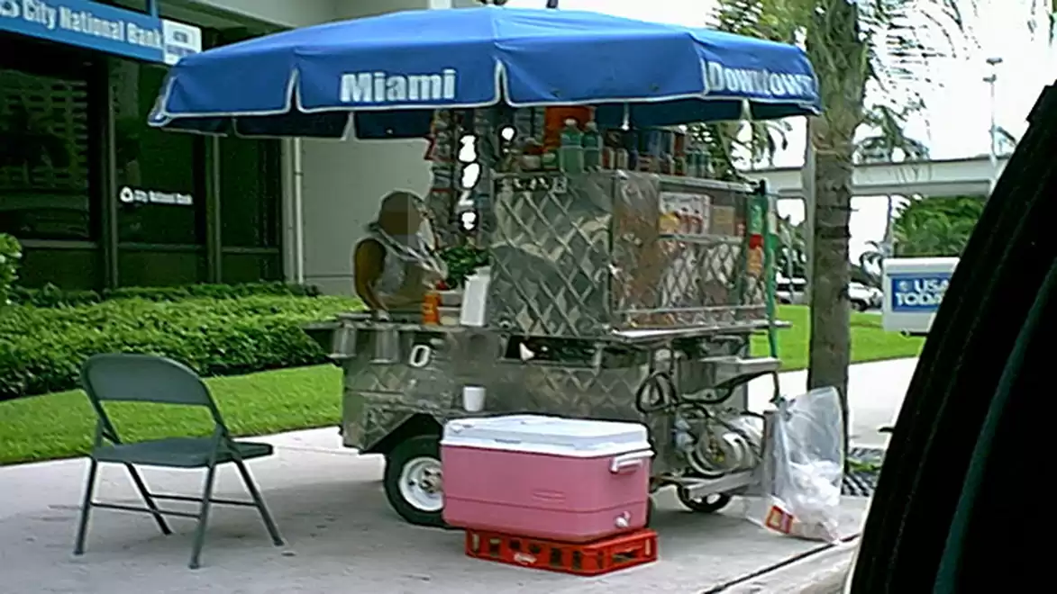 A mobile street vending stand in Miami with a blue umbrella, a chair, and a cooler on the sidewalk.