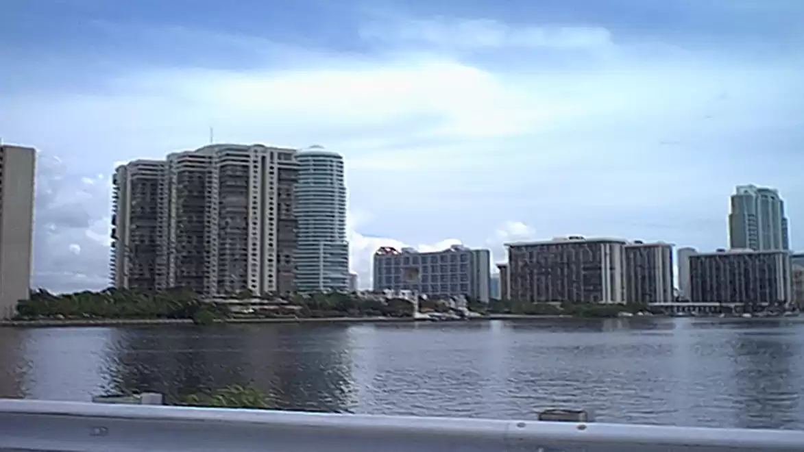 View of modern high-rise buildings on a riverbank under a cloudy sky.