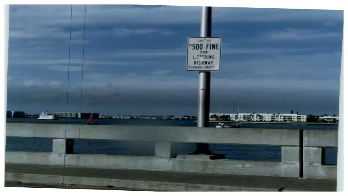 Road sign on a bridge indicating a $500 fine for littering, with a lake and buildings in the background.