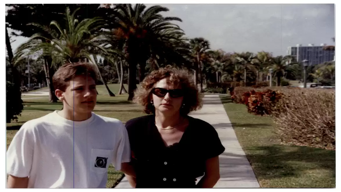 A young man and a woman wearing sunglasses are walking on a sidewalk with palm trees in the background.