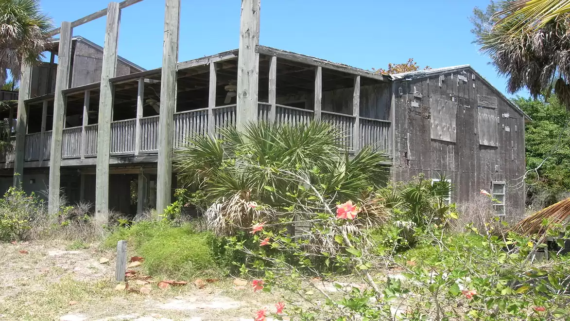 Abandoned wooden building with overgrown vegetation in the foreground on a sunny day.