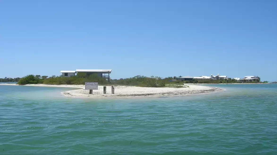 Small sand island in clear blue water with some buildings in the background under a clear blue sky.