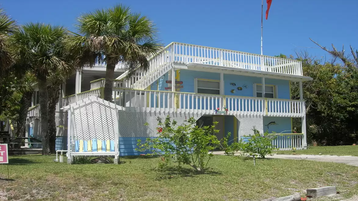A two-story blue building with a porch, surrounded by palm trees and tropical vegetation.