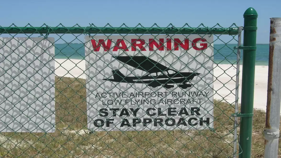 A warning sign on a fence reading: 'Active Airport Runway. Low Flying Aircraft. Stay Clear of Approach.' near the beach.