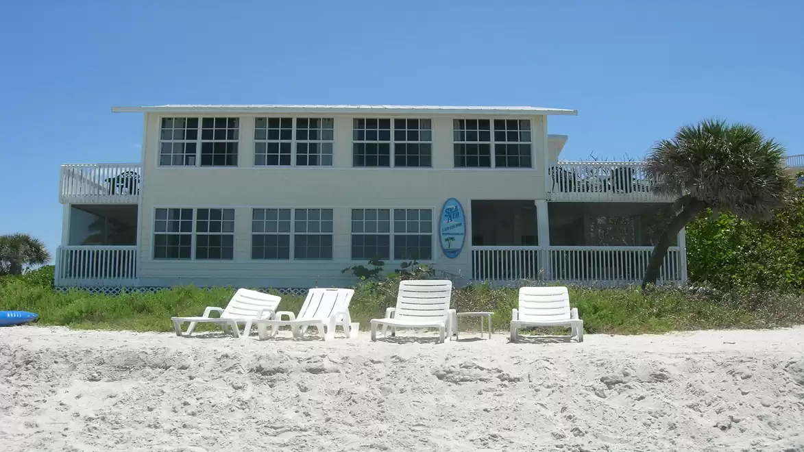 A white beachfront house with a large terrace and several lounge chairs in the sand in front, under a clear blue sky.