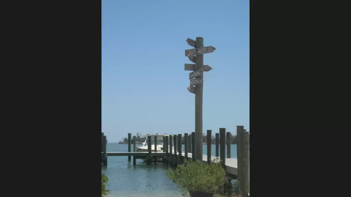 A wooden signpost at a pier pointing toward a scenic coastal view with calm water in the background.